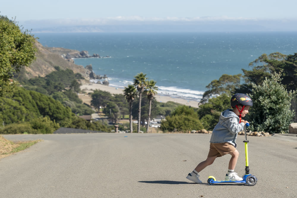 Kid riding a Micro Kickboard scooter on a coastal road above the ocean
