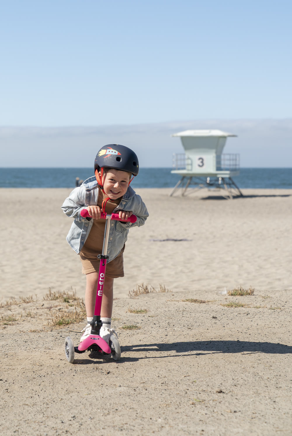 Kid on a pink Micro Kickboard scooter at the beach by a lifeguard tower